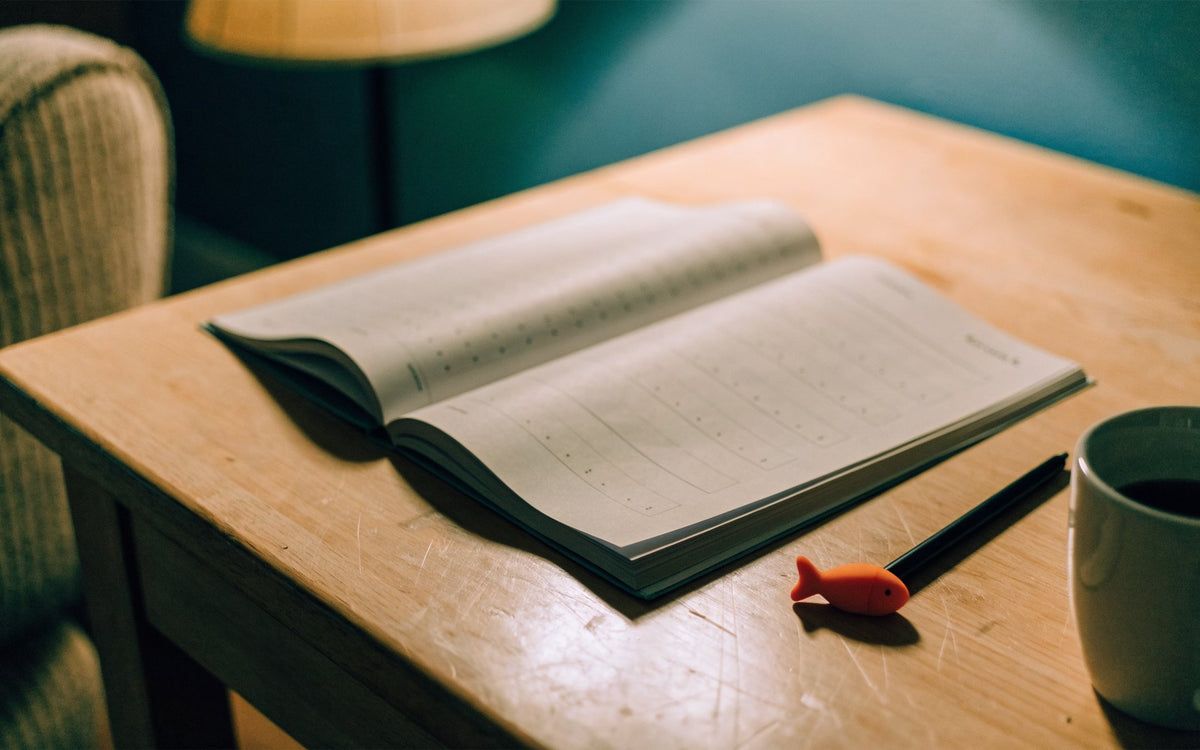 Une table de bois le soir, sous une lampe chaude : un cahier Ecrilo ouvert, le stylo et son guide-doigts orange posés à côté